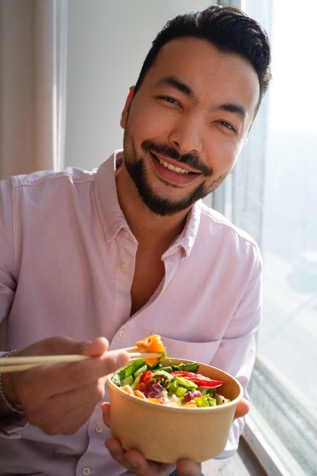 A bearded man with a pink button-down shirt uses chopsticks to pick up a piece of fruit from an Asian salad.
