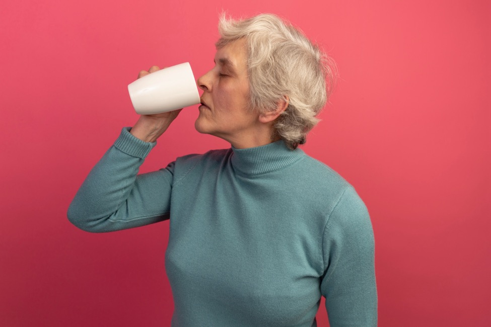 A woman drinking from a white cup against a coral background.